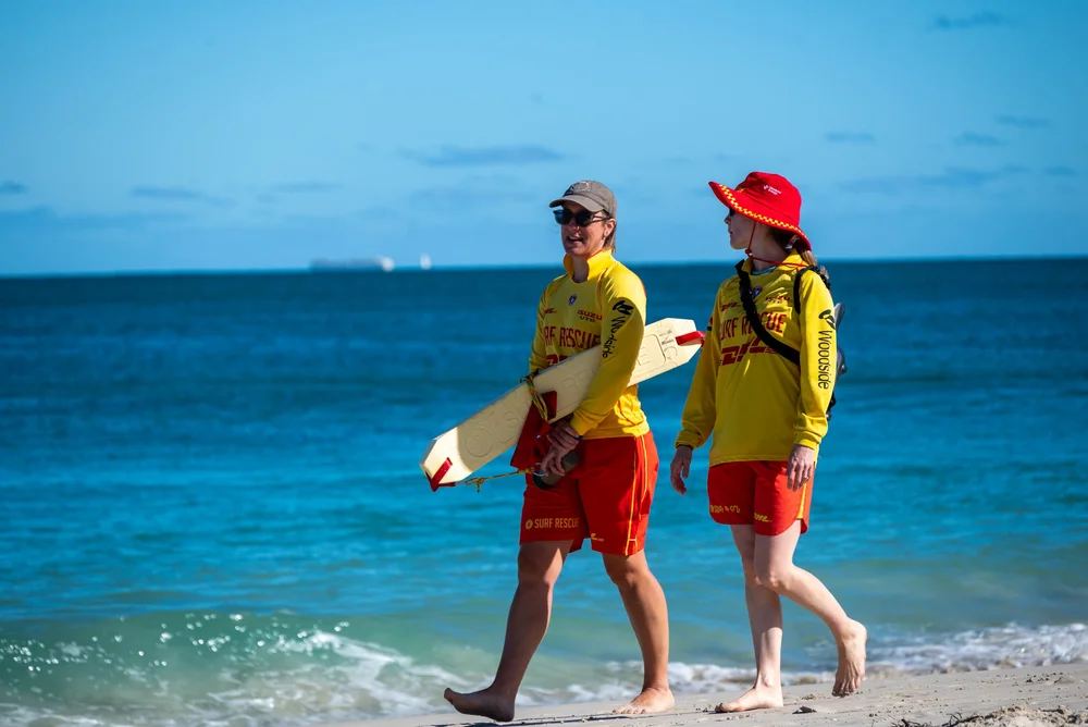Two North Cott patrollers in red and yellow walking the beach with a rescue board